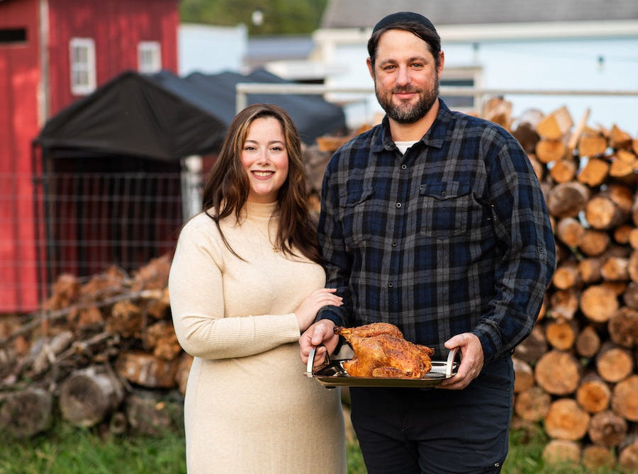 Man and woman holding a roasted turkey in front of a barn and stack of logs.